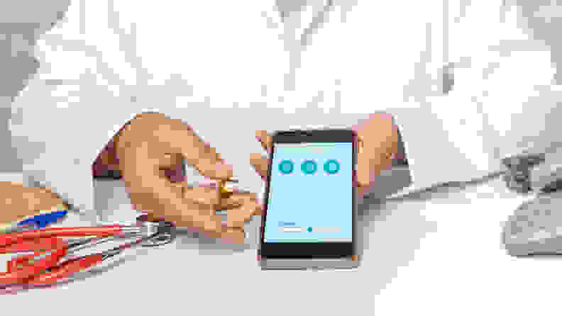 A doctor in a white coat sits at a desk next to a stethoscope while holding a smart phone and a hearing aid in hand.