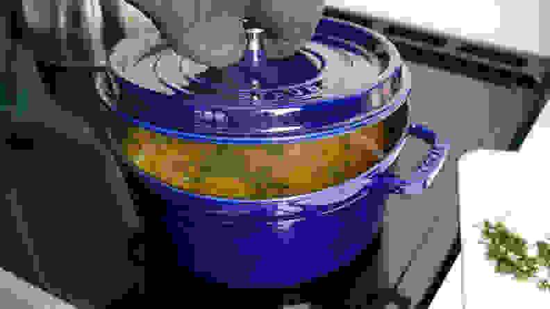 A person lifts the lid off of a blue Staub Dutch oven to reveal chicken soup cooking inside.