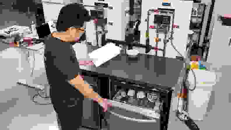 Our Senior Manager of Lab Operations, Jonathan Chan, stands before a dishwasher ready for testing in our labs, running through a checklist on a clipboard. Jonathan opens the dishwasher's door, showing an interior filled with dirty dishes.