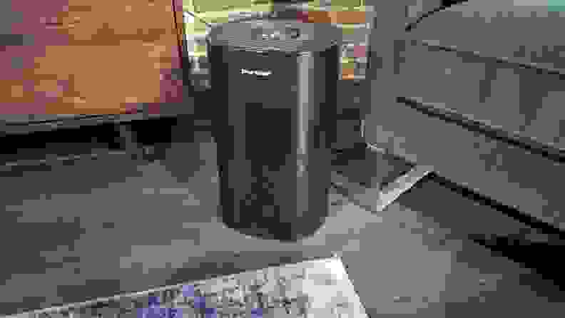 A black  Puro Air 240 air purifier stands aside a gray couch and on a brown wood floor.