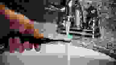 A man rinses the head of a black electric toothbrush under a bathroom faucet.