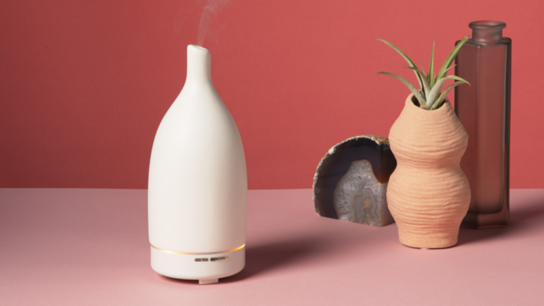 Saje Aroma Om essential oil diffuser on a pink surface with a geode, potted succulent, and a cylindrical glass vase to the right.