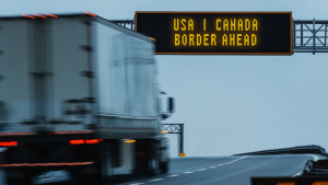 A truck drives a highway under a sign that reads USA Canada border