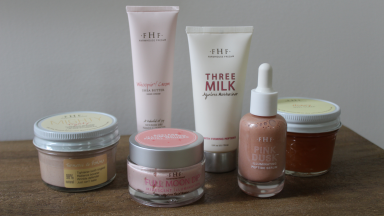 FarmHouse Fresh Mighty Tighty Turmeric & Banana Tightening Mask, Whoopie! Shea Butter Hand Cream, Full Moon Dip, Three Milk Ageless Moisturizer, Pink Dust Illuminating Peptide Serum and Honey Lavender Salt Body Scrub all gathered together on top of wooden surface.